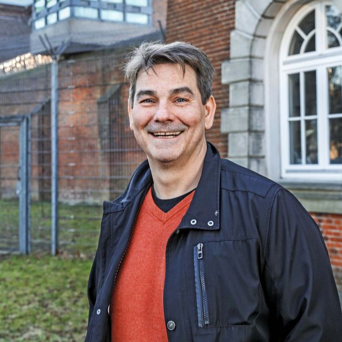 Porträt eines Mannes mit braunen Haaren und lächelndem Gesicht, in einem dunklen Oberteil.